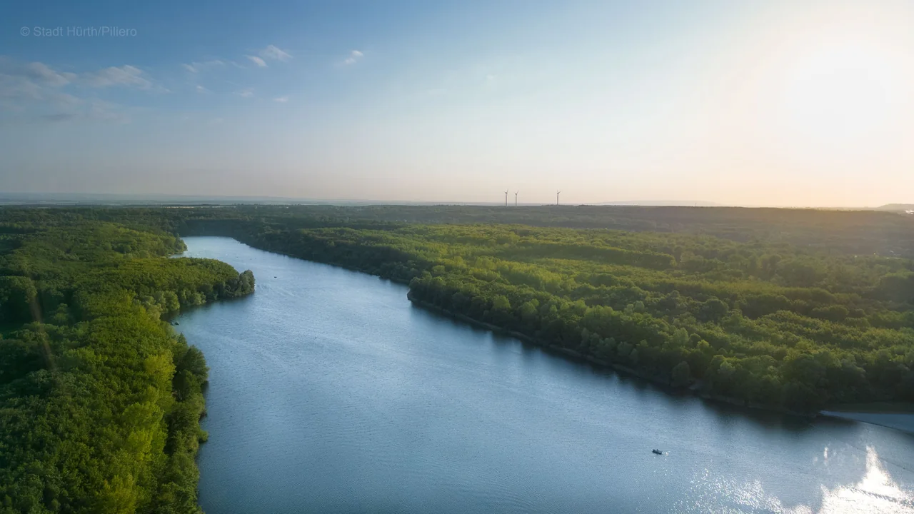 Drohnenaufnahme eines langgestreckten Sees zwischen dichtem Wald im Abendlicht