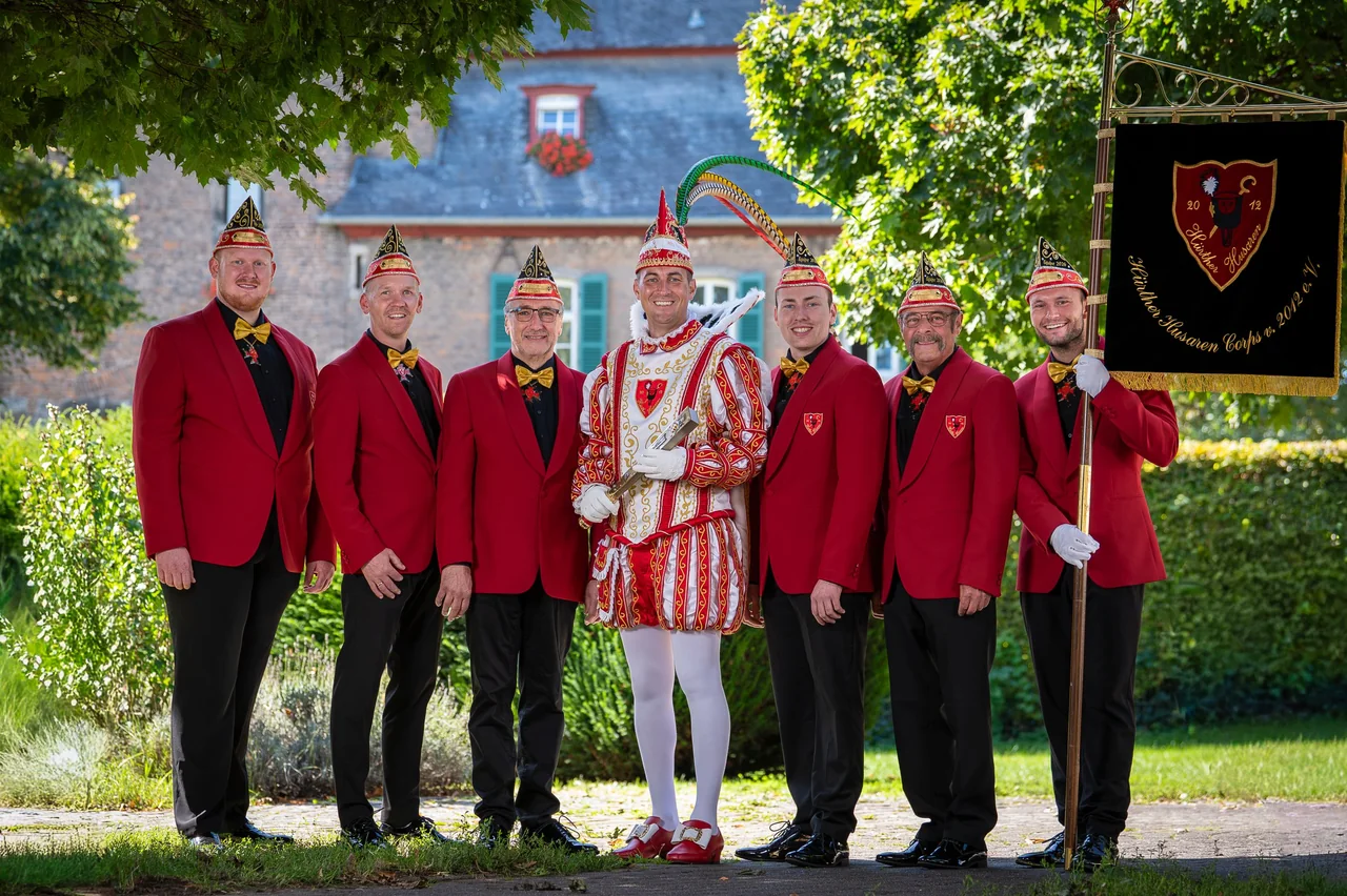 Karnevalsprinz mit seinem Corps in roten Jacken und Standarte vor einer Burg